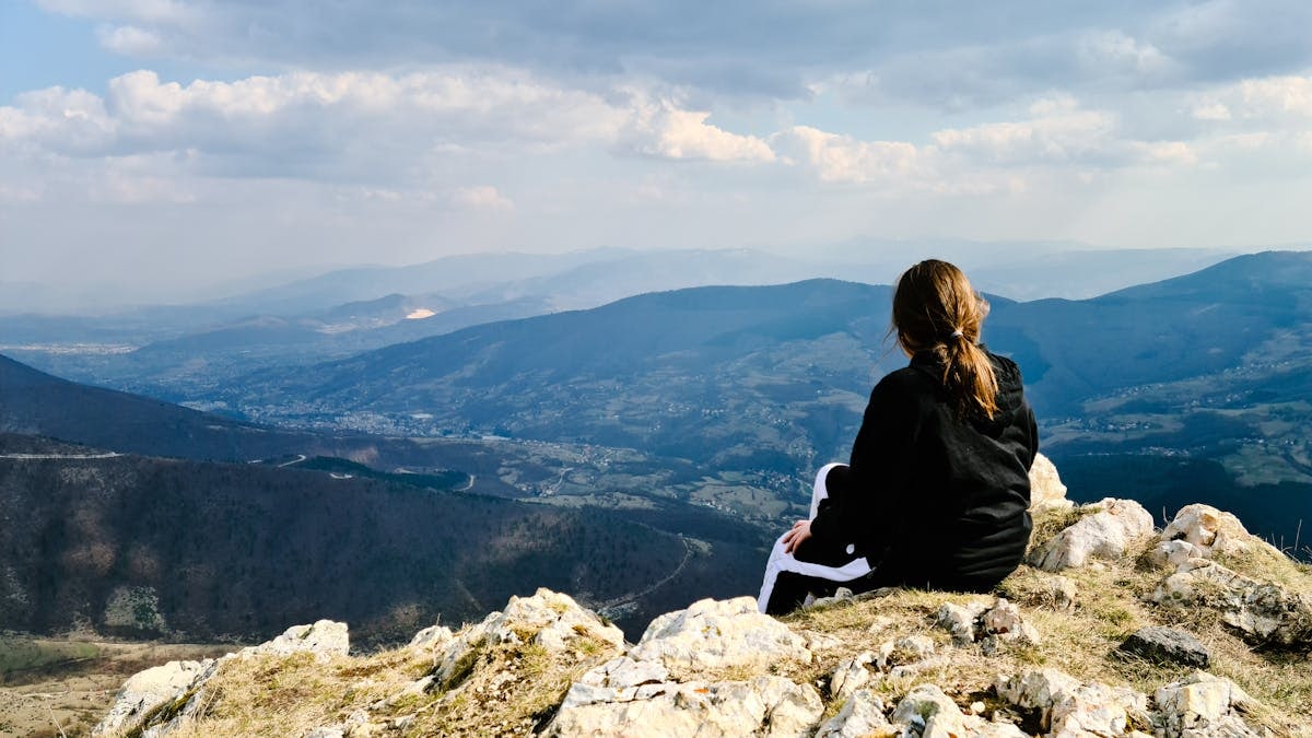 A woman sits on a rocky mountain peak, enjoying a panoramic view of the valley below.